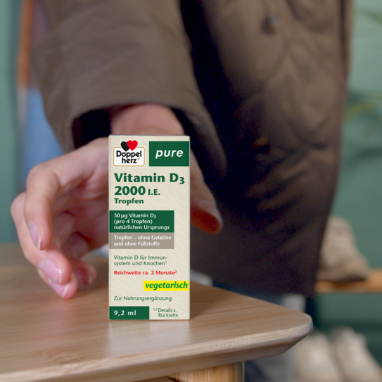 Hand greift nach Doppelherz pure Vitamin D3 2000 I.E. Tropfen auf einem Tisch