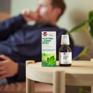 Produkt steht im Vordergrund auf einem Hocker und im Hintergrund hustet ein Mann auf dem Sofa sitzend