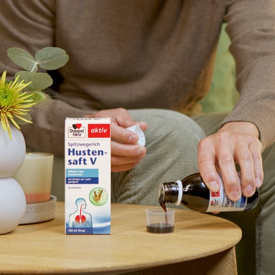 Das Produkt steht auf einem Tisch und eine Hand schenkt den Hustensaft in den Messbecher