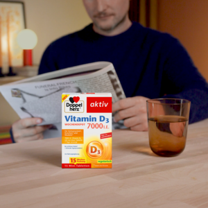 Doppelherz Vitamin D3 7000 I.E. auf Tisch mit Wasserglas, Mann mit Zeitung im Hintergrund