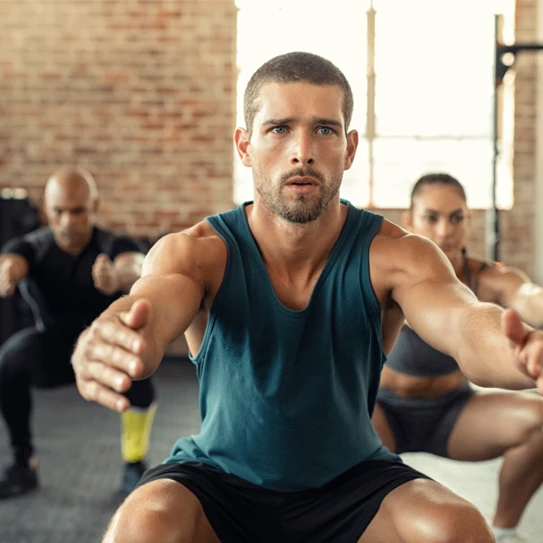 Junger Mann, der mit Kniebeugen im Fitnessstudio mit Menschen im Hintergrund trainiert.