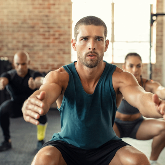Junger Mann, der mit Kniebeugen im Fitnessstudio mit Menschen im Hintergrund trainiert.